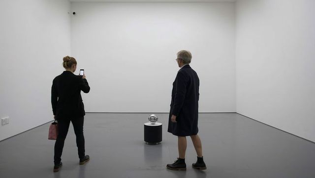Minimalist gallery visitors observing reflective metallic orb sculpture on pedestal