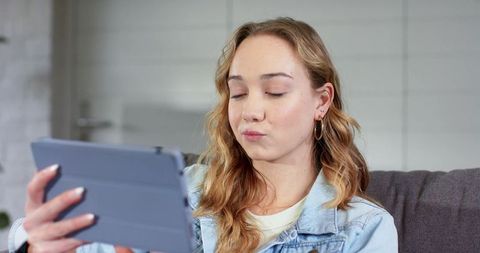 Young woman holding tablet on couch wearing denim jacket and hoop earrings, relaxed lifestyle