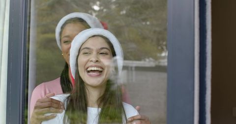 Joyful holiday embrace: mother and daughter laughing through window wearing santa hats