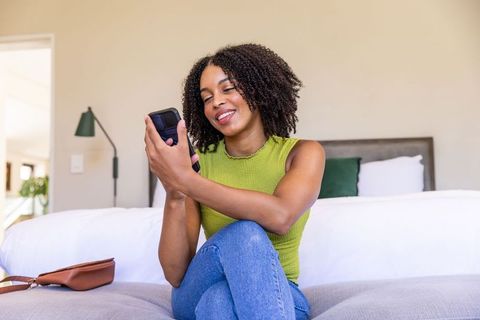 Smiling woman sitting on bed scrolling smartphone in bright modern bedroom interior