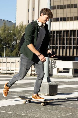 Urban teen skateboarding across rooftop plaza pushing off with smartphone and backpack
