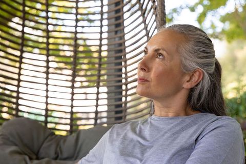 Senior woman relaxing in woven rattan hanging chair on veranda with thoughtful gaze