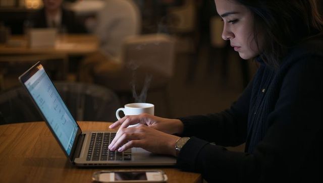 Focused woman typing on laptop in cozy cafe with steaming coffee cup and smartphone
