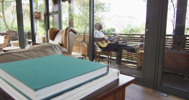 Man sitting on patio playing acoustic guitar and relaxing in a sunny atmosphere. Comfortable indoor space with large windows offering a view of nature-filled backyard. Suitable for themes of relaxation, tranquil lifestyle, music enjoyment, indoor-outdoor living, and cozy home settings.
