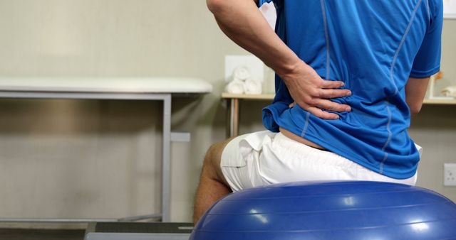 Man sitting on stability ball in physical therapy room, holding lower back in apparent pain, suggesting injury rehabilitation. This image can be used in healthcare blogs, physical therapy advertisements, medical websites, or fitness guides on back injury exercises.