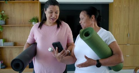 African american women checking workout app and holding yoga mats in living room
