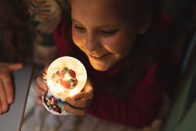 Caucasian girl holding a glowing christmas globe close to her face while smiling inside a blanket fort. beside her is her mother.
