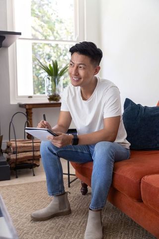 Asian man sitting on orange sofa smiling and writing in spiral notebook wearing smartwatch