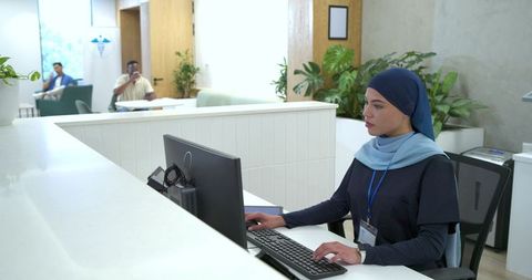 Receptionist Wearing Hijab Typing at Modern Clinic Front Desk, Welcoming Visitors