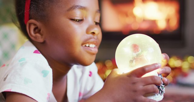 Child enjoying the warmth of a glowing snow globe by the fireplace. Ideal for holiday, winter, Christmas themes in advertisements, blog posts, and greeting cards.