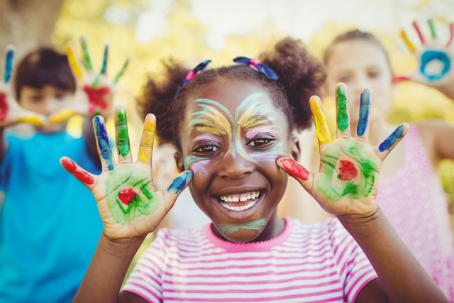 This image captures a joyful girl with face paint showing her colorful painted hands while smiling in a park. Ideal for use in advertisements, educational materials, and websites promoting children's activities, creativity, and outdoor fun. Perfect for illustrating themes of happiness, friendship, and artistic expression.