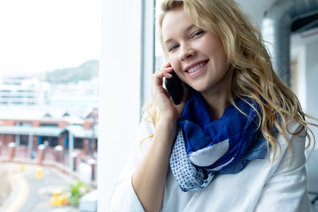 Front view of beautiful young caucasian businesswoman looking at camera while talking on mobile phone near window in a modern office