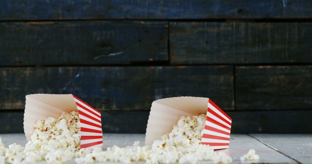 Two red and white striped popcorn boxes are tipped over with popcorn spilling out onto a wooden floor. This image is perfect for promoting movie nights, cinema-related events, as well as snack foods. It can also be used in marketing materials for theaters, casual dining establishments, or family entertainment spaces.