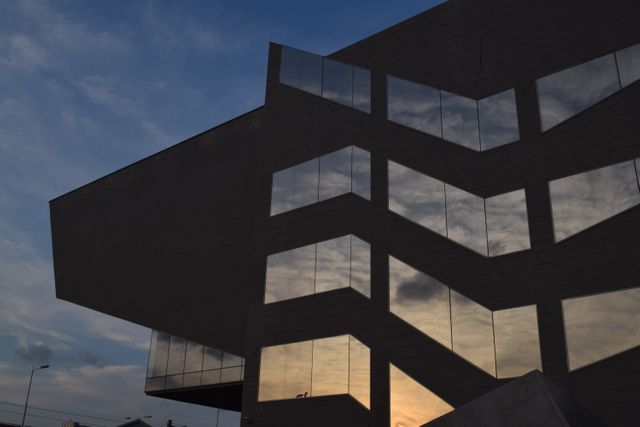 The image captures a sleek and modern building facade with large glass windows reflecting the colors of the sunset. The unique geometric shapes of the structure create an interesting visual contrast with the soft evening sky, making it ideal for themes related to contemporary architecture, urban landscapes, and architectural design. This can be used in architectural magazines, urban planning articles, or advertisements for real estate developments.