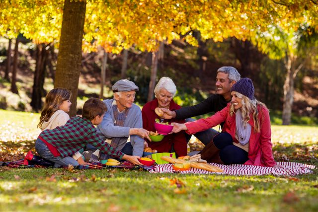 Multi-generation family enjoying a picnic in an autumn park. Grandparents, parents, and children are sitting on a blanket, sharing food and smiling. The vibrant fall foliage creates a warm and inviting atmosphere. Ideal for use in advertisements, family-oriented content, and articles about family bonding, outdoor activities, and seasonal events.