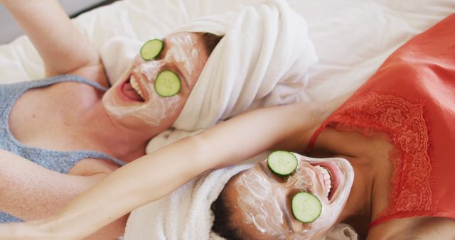 Diverse happy female friends with towels on heads cleansing masks and cucumber on eyes lying on bed. female friends hanging out enjoying leisure time together.