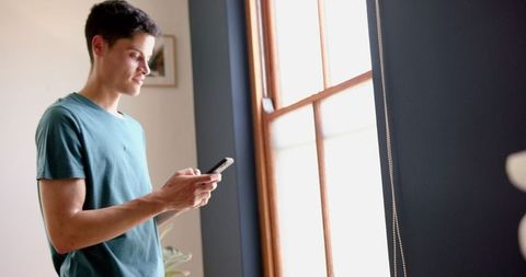 Young Man Using Smartphone Standing by Sunny Window at Home