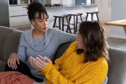 Diverse female friends sharing honest conversation on gray sofa in cozy open-plan home