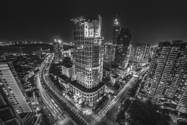 Monochrome photo of a city at night showcasing tall skyscrapers and bustling streets with light trails. Suitable for use in urban development materials, architectural studies, and as artwork for modern wallpapers or city-themed presentations.