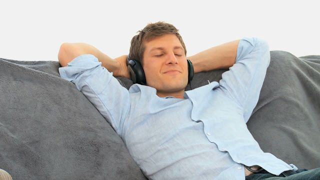 Young man wearing headphones, lying back on a sofa with hands behind his head, appearing relaxed and content. Suitable for promoting leisure time, mental health, and the benefits of music. Works well for lifestyle blogs, relaxation tips, and advertisements related to home comfort or entertainment products.