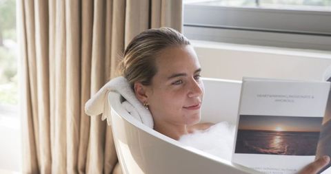 Relaxing woman reading in bubble bath by sunlit window, towel neck support, calm morning