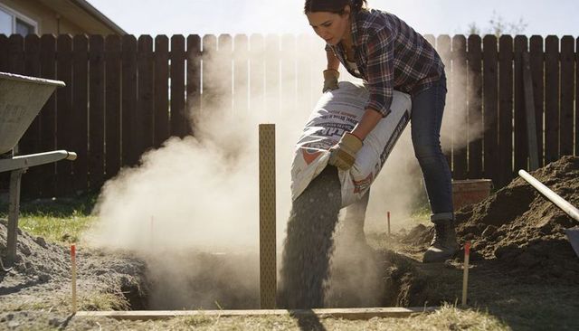 Woman pouring gravel into yard trench for drainage while wearing flannel, gloves and boots