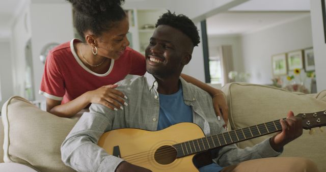 A joyful couple in a cozy home environment playing guitar and smiling at each other in a spacious living room. Suitable for depicting themes of music, relationship, leisure time, and a happy home life. Perfect for websites, advertisements, blogs related to music, home lifestyle, and relationships.