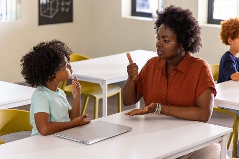 African american teacher teaching sign language to child in bright classroom with laptop