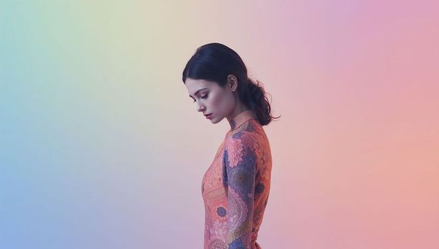 Serene woman standing in profile bowing head wearing patterned dress on pastel gradient