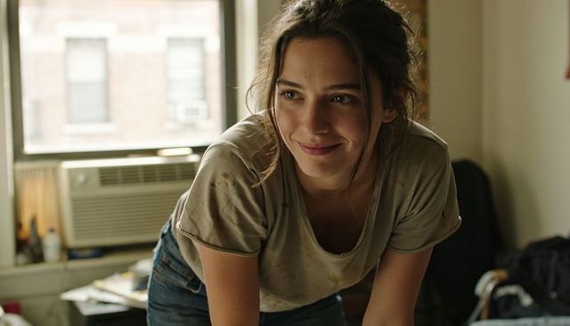 Smiling woman leaning forward in sunlit apartment wearing stained t-shirt and jeans