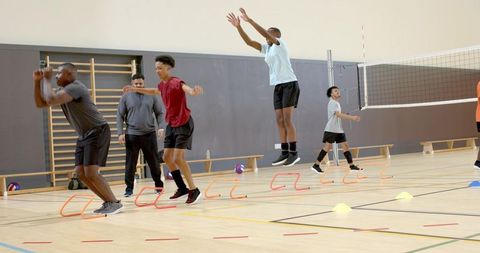 Diverse male athletes practicing agility drills with low hurdles in indoor volleyball gym