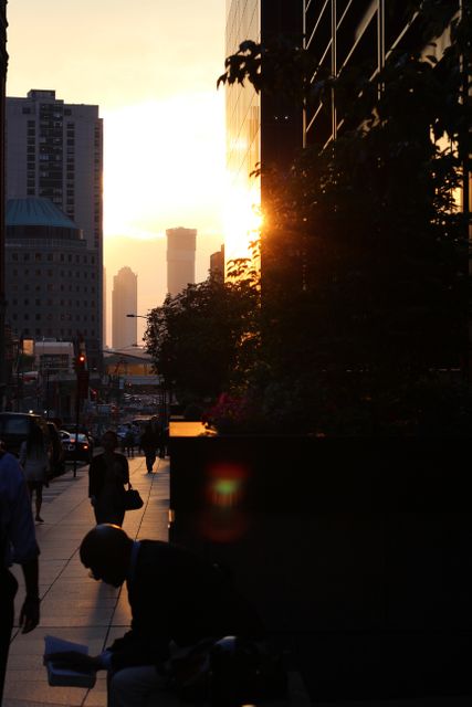 People are walking along a busy city street at twilight with the sun setting, casting silhouettes of skyscrapers and high-rise buildings. This stock photo is perfect for projects related to urban lifestyle, business districts, evening commutes, or city life. It conveys a sense of modernity, hustle, and the end-of-day atmosphere in a metropolitan area.