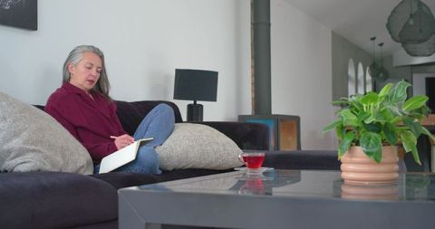 Senior woman writing in journal on sofa at home with tea, indoor plant and cozy stove