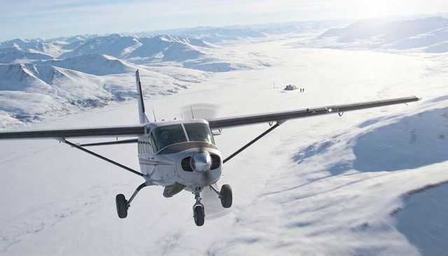 Single-engine high-wing aircraft flying low over snowfield toward remote mountain hut