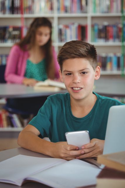 Schoolboy smiling while using mobile phone in library, surrounded by books and study materials. Ideal for educational content, technology in education, student life, and academic resources.