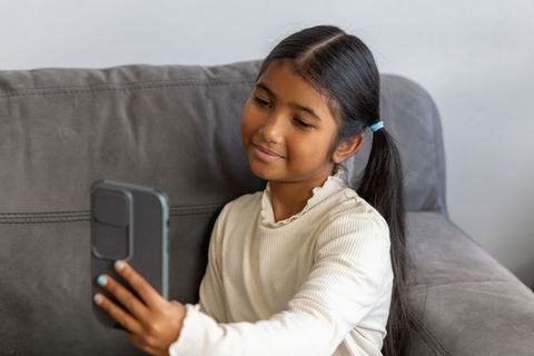 Indian girl taking selfie on smartphone while sitting on gray sofa at home