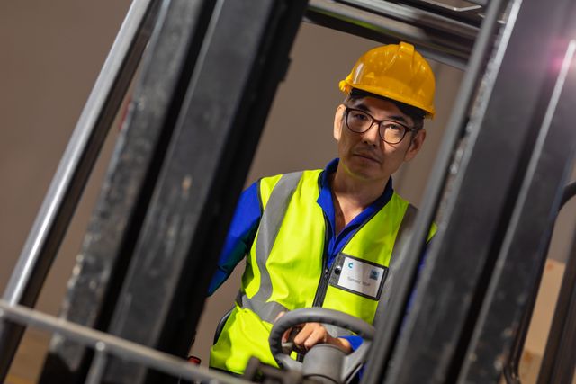Portrait of asian mature male warehouse worker wearing eyeglasses and hardhat driving forklift. truck, unaltered, warehouse, logistics and shipping occupation.