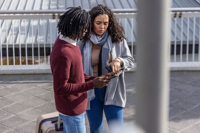 Diverse young travelers checking phone on urban transit platform with carry-on luggage