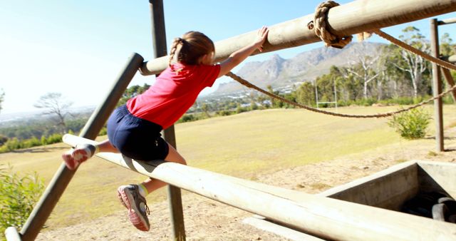 Child navigating an obstacle course in a sunlit outdoor setting. Perseverance and concentration visible as they hold onto the structure. Suitable for advertisements or articles promoting children's outdoor activities, creating a narrative around physical development, or marketing outdoor gyms and playgrounds.