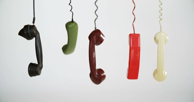 Vintage telephone handsets in black, green, red, and cream colors are suspended against a white background, with copy space. Their twisted cords add a dynamic element, evoking a sense of communication from a bygone era.