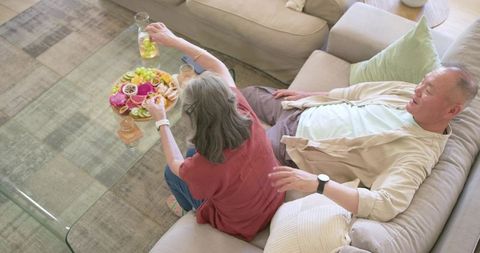 Senior couple snacking on colorful fruit platter on sofa, relaxing at home