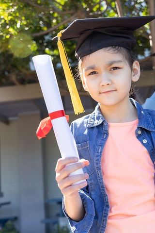 Asian child graduate holding diploma wearing mortarboard with yellow tassel in sunlit courtyard