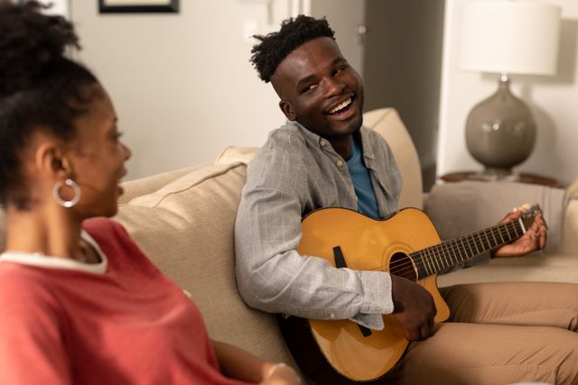 Happy african american young man singing and playing guitar while looking at girlfriend on sofa. hobbies, unaltered, lifestyle, home, love and togetherness concept.