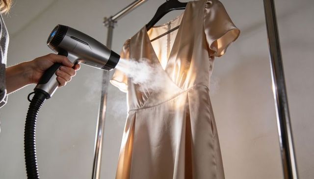 Steaming champagne satin dress on chrome rack with handheld steamer preparing elegant gown