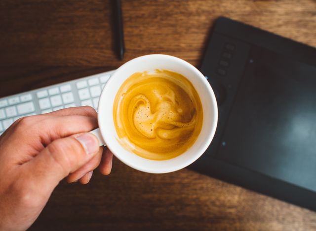 A hand is holding a coffee mug filled with coffee above a work desk that includes a keyboard and a graphics tablet. This is ideal for themes involving technology, workspace, office environment, remote work, and startup culture. It can be used in articles or advertisements about modern working conditions, productivity tips, and morning routines.
