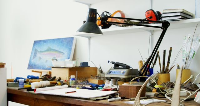 Interior of an artisan's workshop featuring a cluttered workbench with various tools, a desk lamp, and a painting in the background. Ideal for illustrating craftsmanship, creativity, and artisanal activities. Suitable for articles and content on DIY projects, artistry, and home workshops.