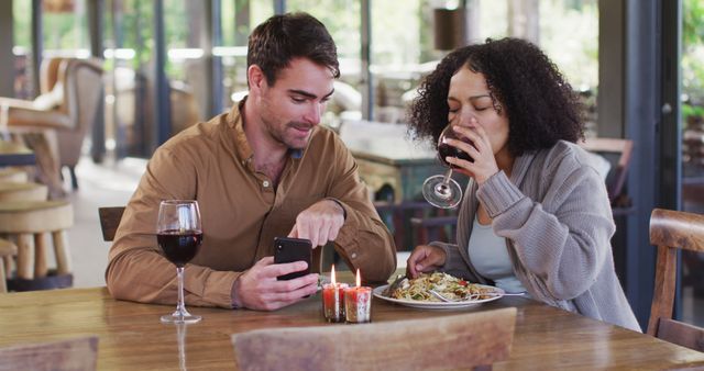 Couple enjoying lunch at a cozy restaurant. Man using smartphone while woman drinks wine and eats. Suitable for concepts related to dating, dining experiences, restaurant settings, relaxation, and enjoying special moments.