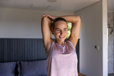Woman stretching arms behind head in bedroom morning routine wearing pink tank top