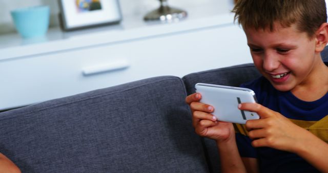A boy is smiling while playing on a tablet, sitting on a gray sofa in a modern living room. This image captures the joy and engagement of children with digital devices. Ideal for use in advertisements for technology products, child-friendly apps, educational content, and home settings. It can also be fit for articles or blogs about children's screen time, modern parenting, or tech-savvy kids.