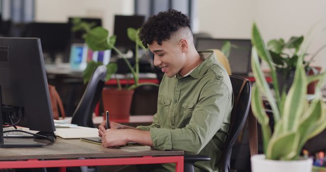 Young professional man taking notes at workspace desk, surrounded by plants, creating a focused and modern work environment. Ideal for business, productivity, office settings, coworking spaces, and modern workplace concepts. Can be used to emphasize concentration, organization, and modern office layouts.